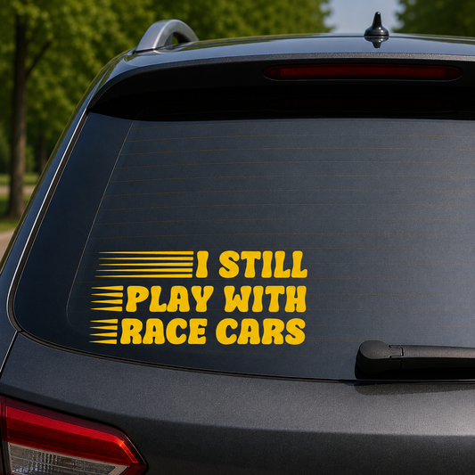 A yellow vinyl decal with the text 'I STILL PLAY WITH RACE CARS' and horizontal racing stripes, shown applied to the back window of a dark gray car.